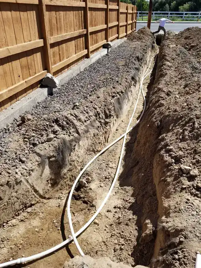 Open trench along a cedar fence with a new white water line laid in the bottom